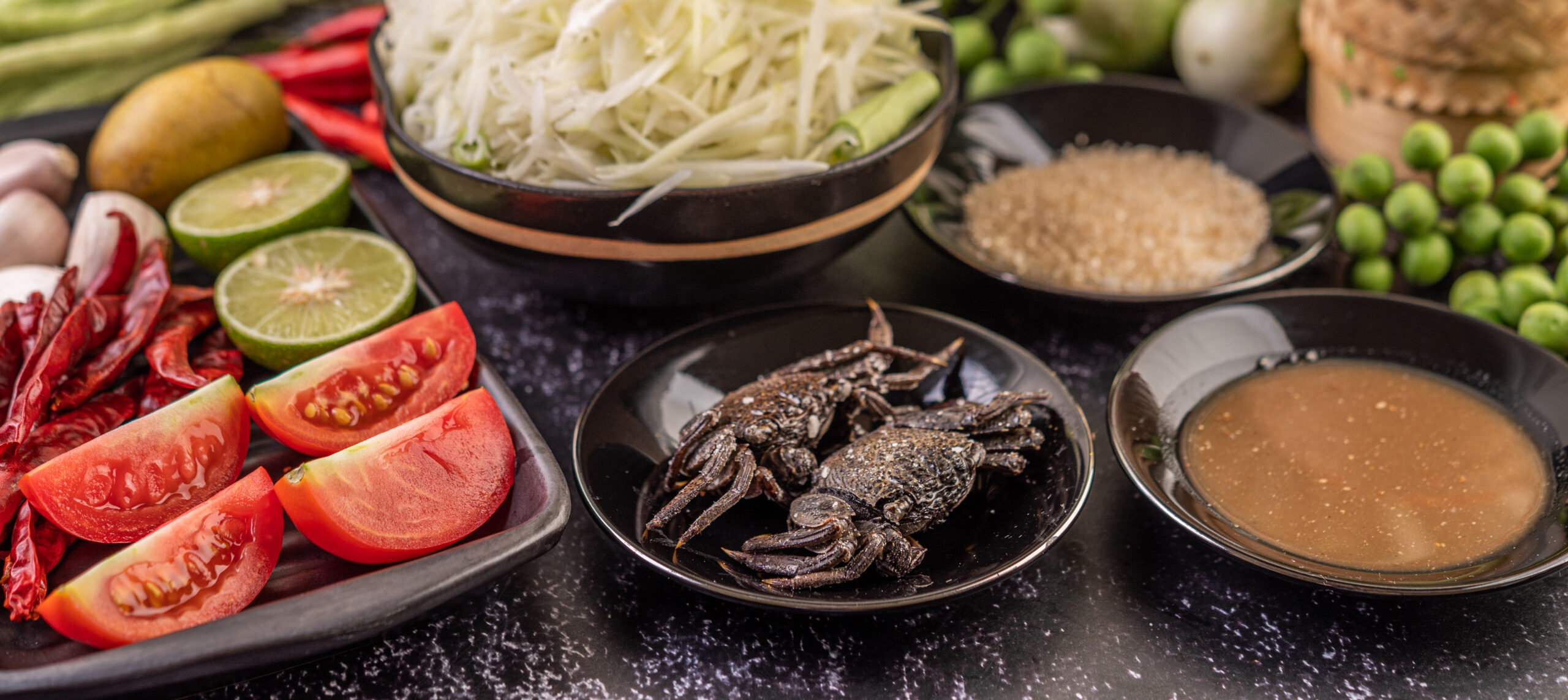 La foto mostra un tavolo con vassoi contenenti pomodori, limoni, insalata di papaya e, al centro, un piatto con degli insetti commestibili